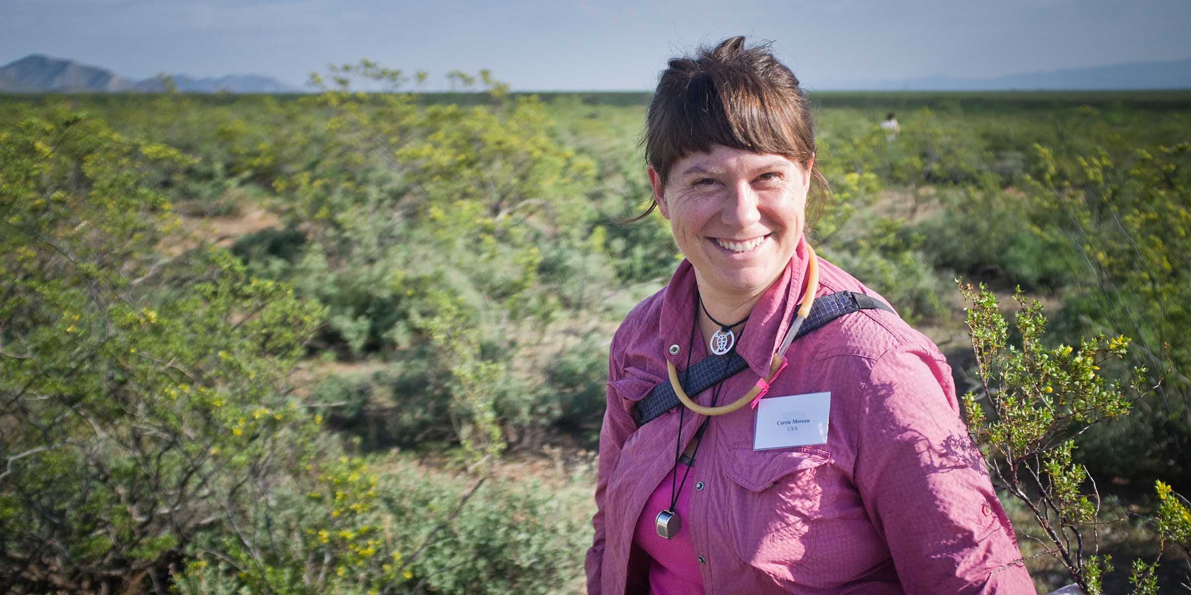 A woman smiling at the camera, standing in a green field with mountains in the background