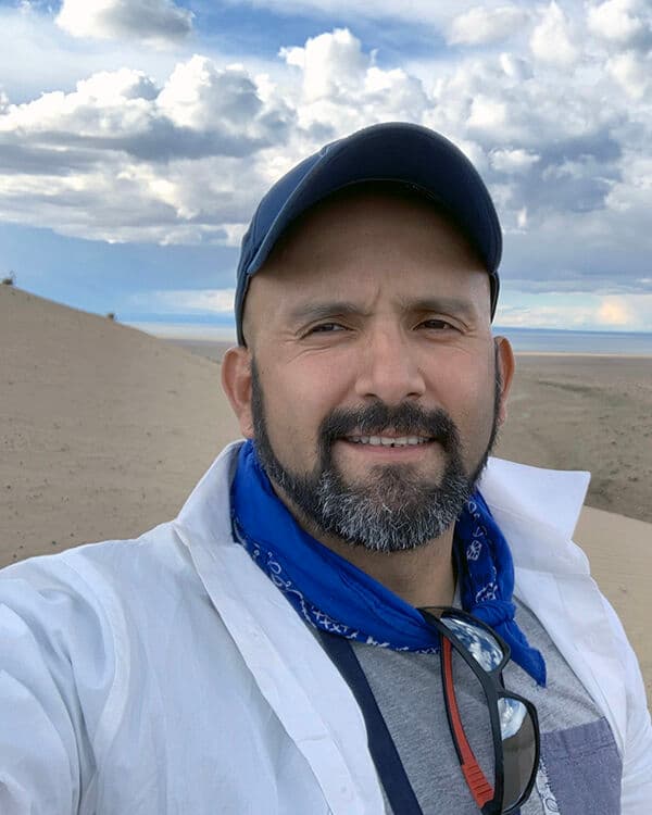 A man in a baseball hat and white coat stands in front of sandy hills and white clouds