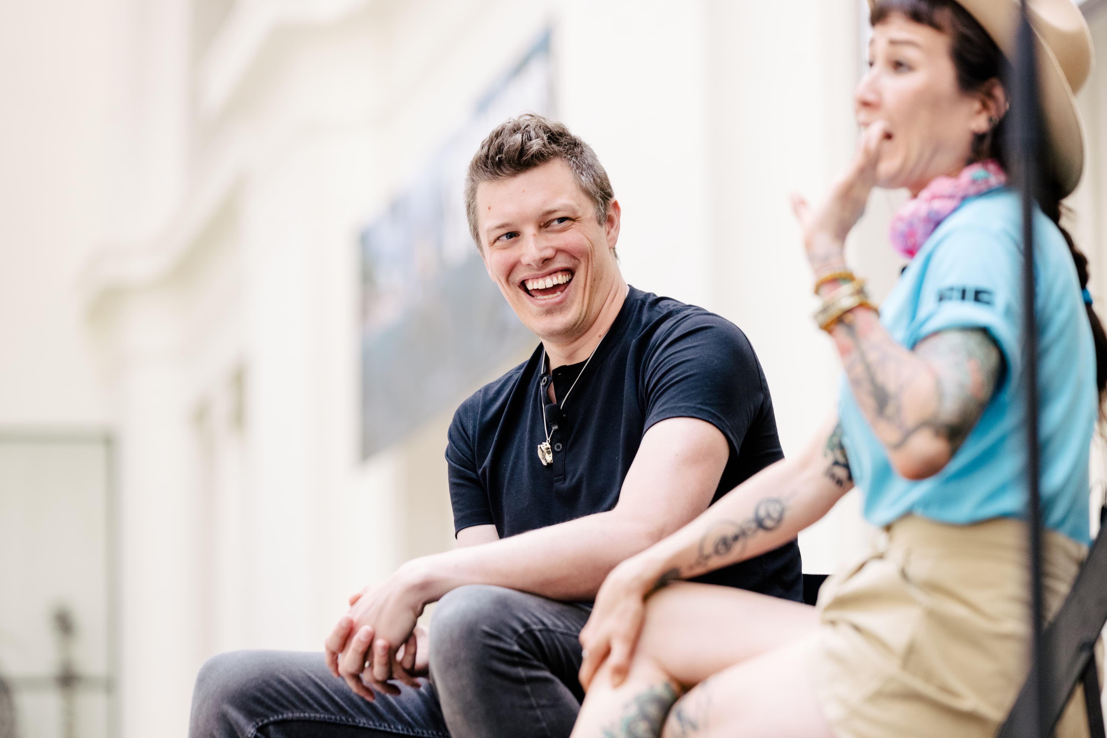 A man wearing a black shirt, smiling as he looks at a woman speaking.