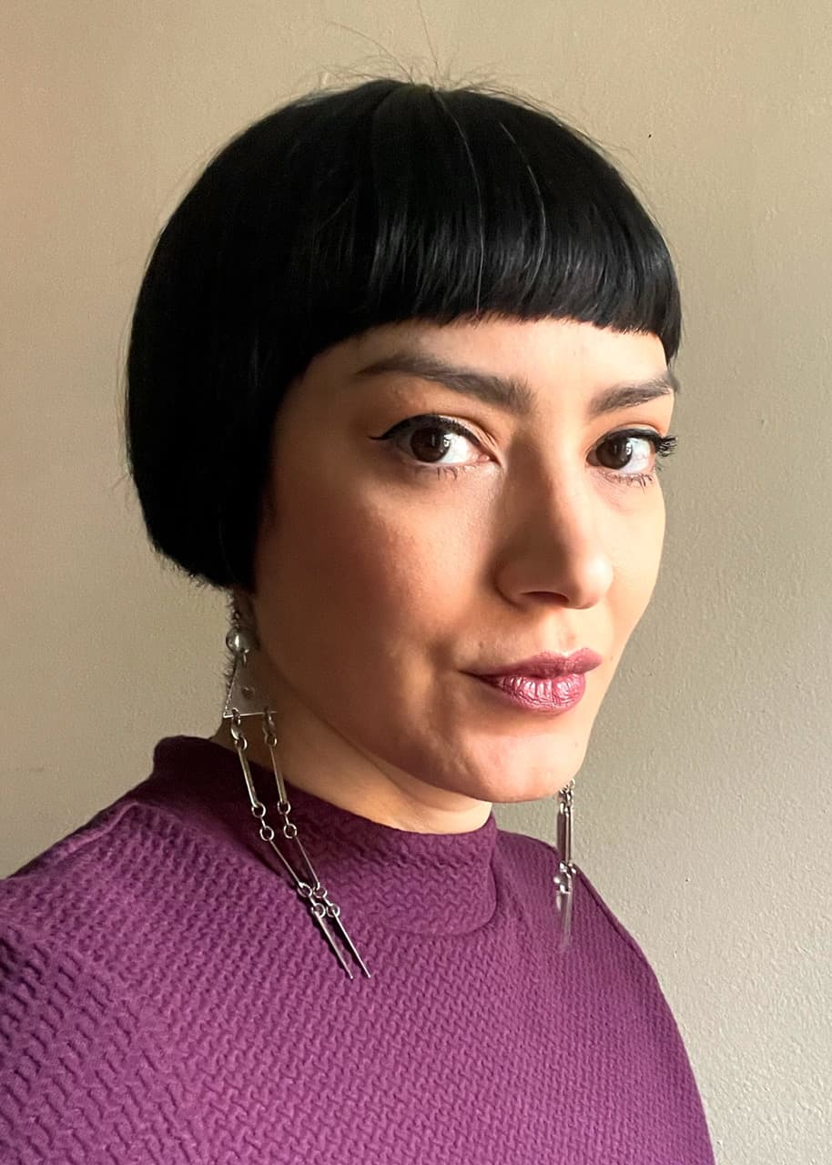 A woman with short black hair, wearing a purple top and silver dangling earrings.