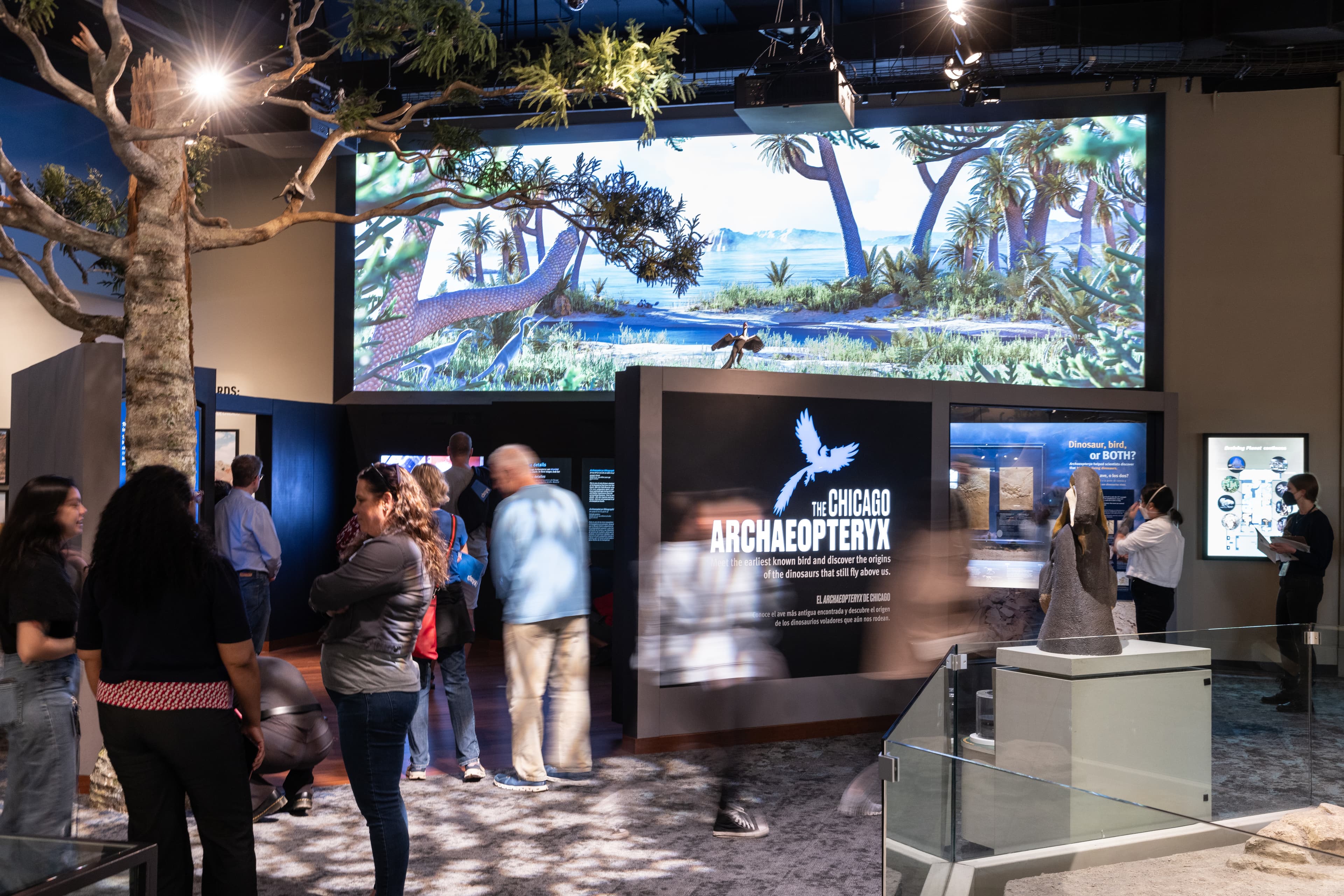 Visitors milling around a museum exhibition, showing a large video screen and other multi-media