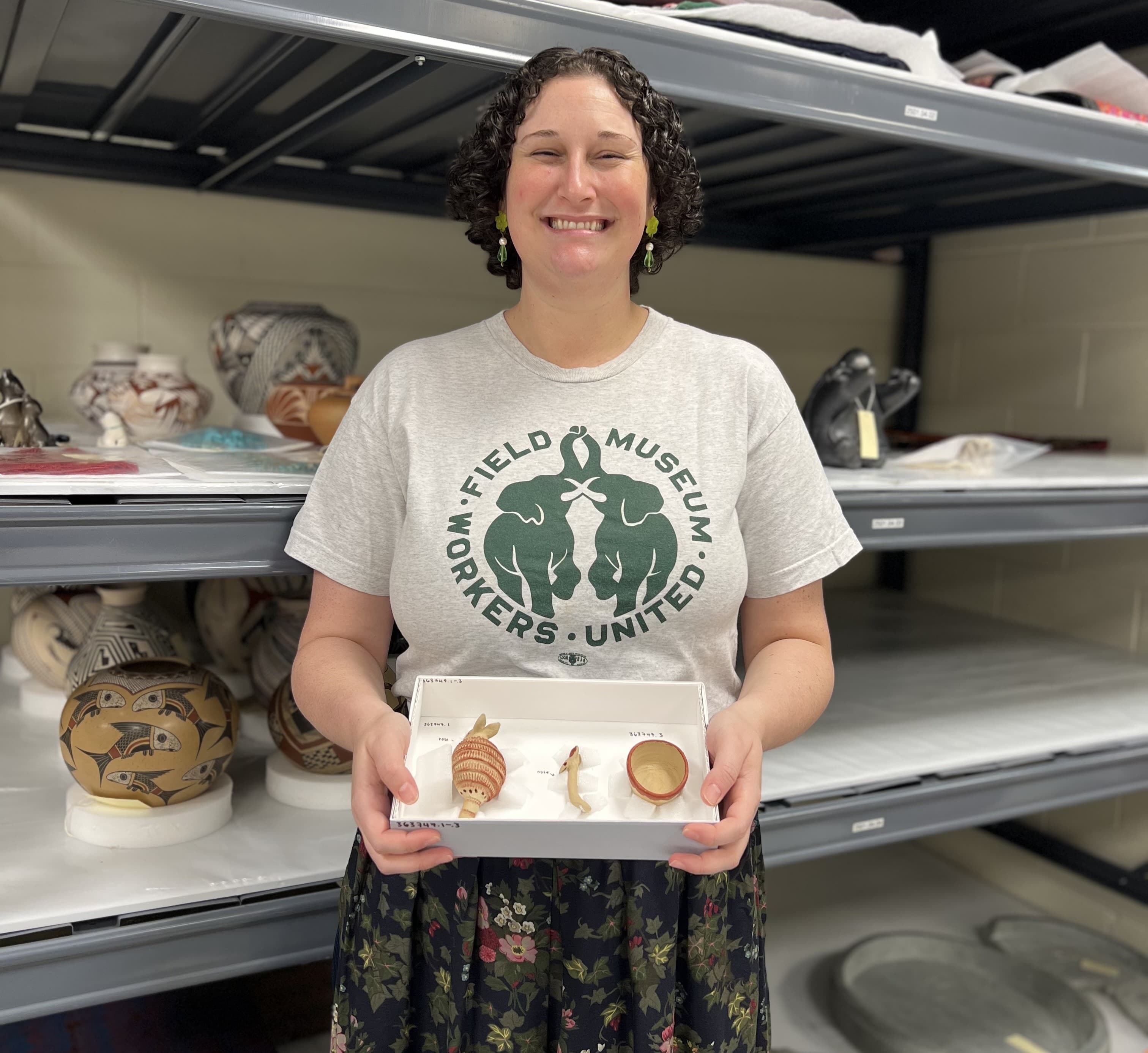 A woman wearing a t-shirt holds a box with three museum specimens as she stands in front of shelves.