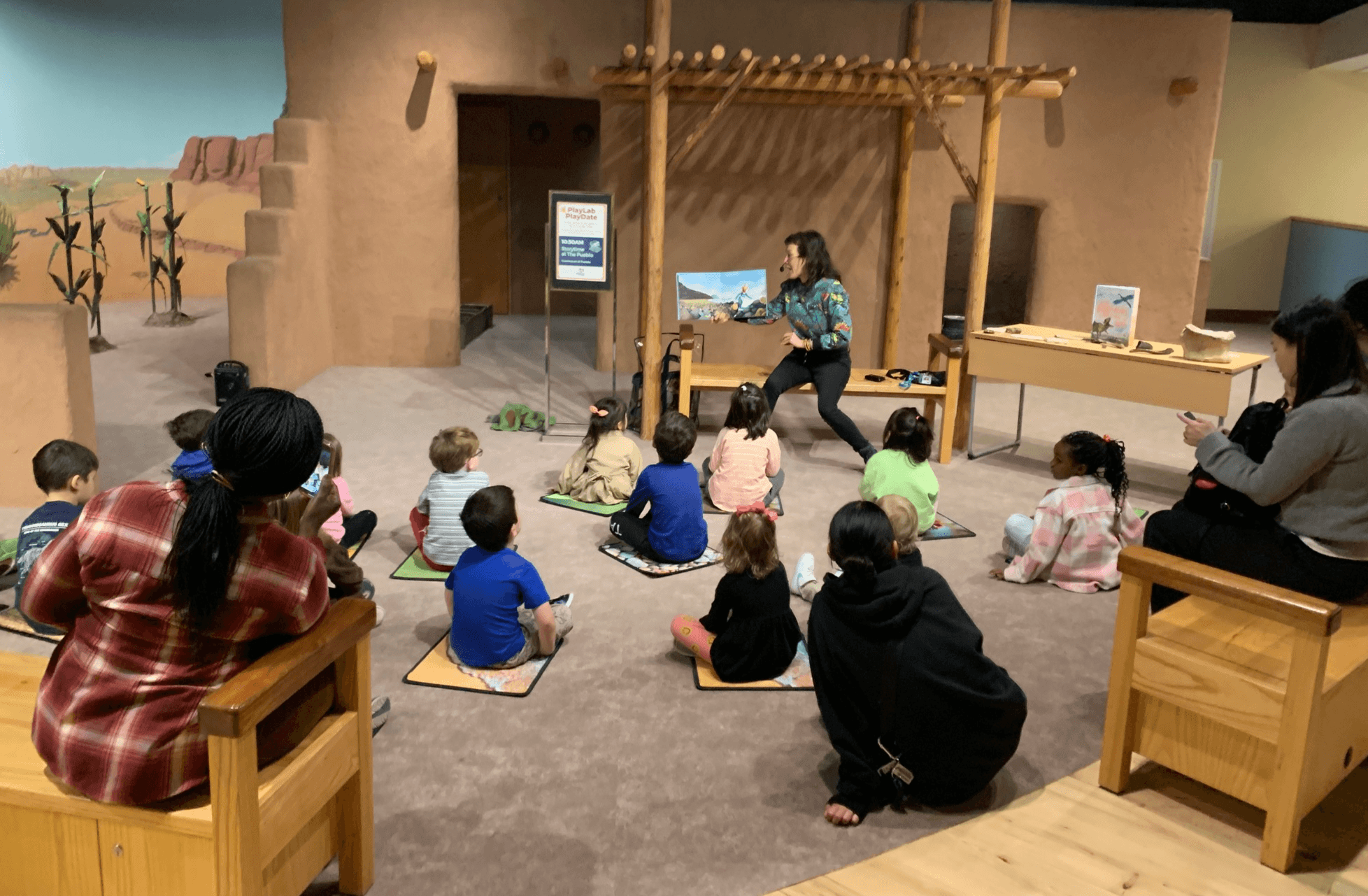 A group of children sit on mats on the floor as a woman reads to them from a picture book. Several other adults are seated on benches around the room.