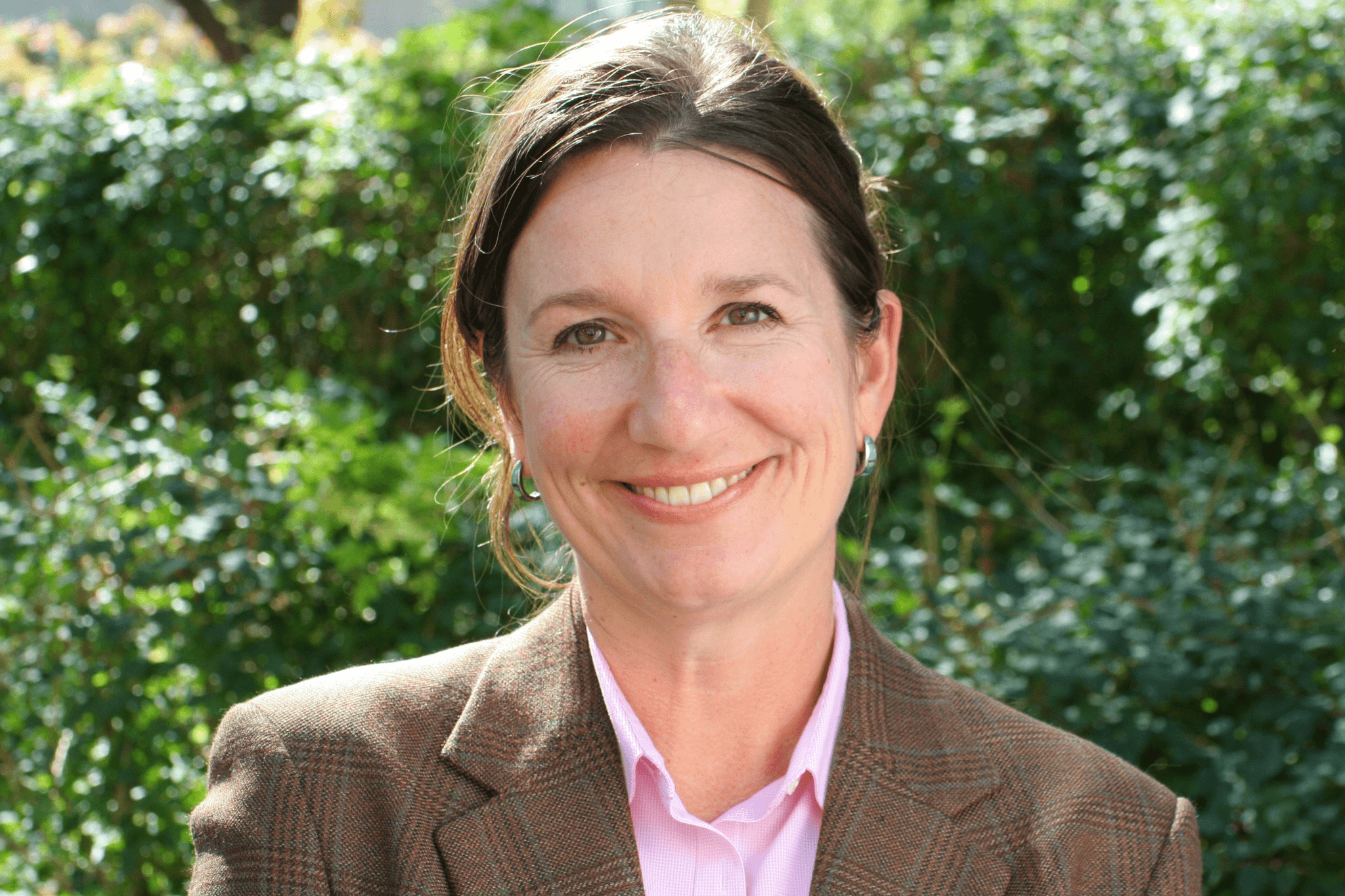 A woman, smiling, wearing a brown plaid jacket and pink shirt. She is standing in front of green shrubs.