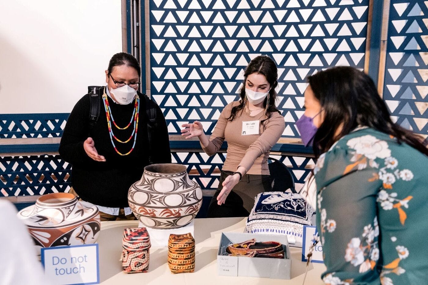 Three people stand around a table motioning to each other over a table with cultural materials displayed, including pottery, baskets, and beaded items.